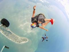 A skydiving cameraman wearing a Red Bull helmet captures another skydiver with an island in the background.