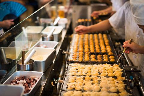 A close-up shot of chefs preparing takoyaki, a popular Japanese street food, on a griddle with visible ingredients like octopus, under bright lighting.
