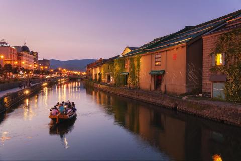 Otaru Holiday House Restoration near Historic Canal/Brewery A boat filled with people cruises down a canal in Otaru, Japan, at twilight, with illuminated buildings and their reflections lining the waterway.