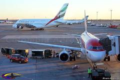 This photograph captures a bustling airport scene featuring multiple airplanes, including an Emirates A380 and a Viva Air aircraft at the gate, surrounded by ground crew and equipment.