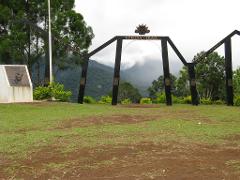 A view of the Kokoda Trail monument on the Sogeri Plateau, featuring black metal structures with inscriptions, a white concrete monument, and a scenic backdrop of mountains and cloudy sky.