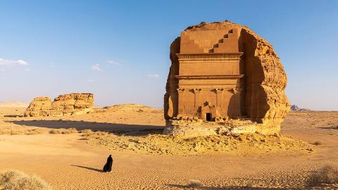A lone figure in black walks across a sandy desert landscape, towards a large, carved rock structure.