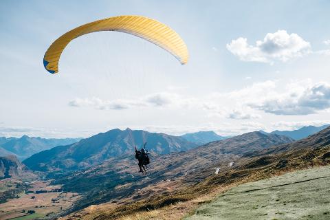 A tandem paraglider soars over a mountainous landscape with a grassy foreground and a partially cloudy sky.