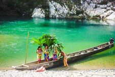 Darien Gap, Rainforest, and Culture from Panama Three children stand near a dugout canoe on the sandy shore of a turquoise river, partially obscured by palm fronds, with rocky formations in the background.