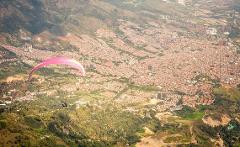 A paraglider soars over a lush, mountainous landscape near a densely populated city.