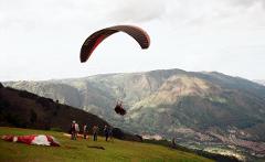 A paraglider soars over a mountainous landscape, with a group of people gathered on a grassy hill below.