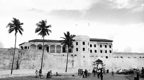 A monochrome view of a historic fort with palm trees, featuring a bustling crowd and parked cars.