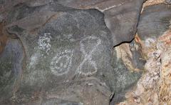 A close-up shot reveals ancient rock art on a cave wall, featuring a spiral and hourglass-shaped symbol created with white pigment.