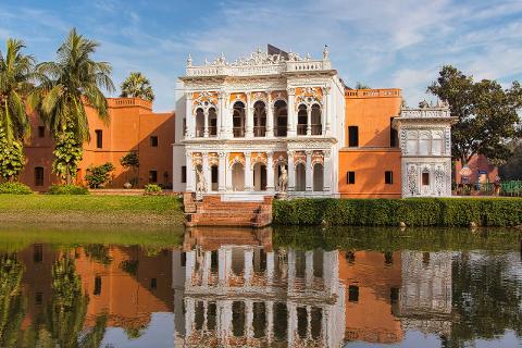 A vibrant palace with intricate designs stands beside a reflective pool, showcasing its stunning architecture under a clear, partly cloudy sky.