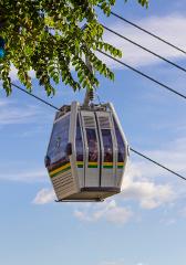 A cable car hangs suspended in the air against a blue sky with wispy clouds, framed by green leaves in the foreground.