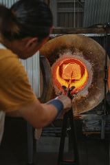 A glassblower uses tongs to extract molten glass from a fiery furnace, possibly crafting chopstick rests or Tonbodama beads at a Tokyo workshop.