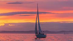 A sailboat glides on a serene sea at sunset, bathed in warm hues of orange and pink, as mountains fade in the distance.