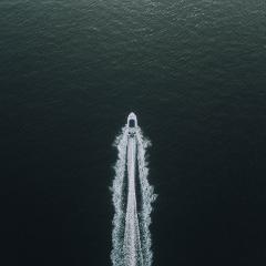 An aerial view shows a white boat speeding through dark, slightly rippled water, leaving a foamy wake behind it.