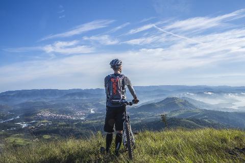 A cyclist with a backpack gazes at the picturesque landscape of misty mountains and a town below from a grassy hilltop under a blue, partly cloudy sky.
