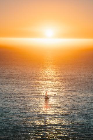 A sailboat glides on the water under a fiery orange sunset, with the sun reflecting on the sea's surface.