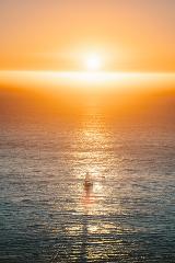 A sailboat glides on the water under a fiery orange sunset, with the sun reflecting on the sea's surface.