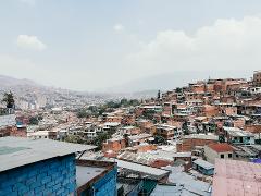 A cityscape view shows a dense, sprawling settlement of closely packed buildings climbing a hillside towards a hazy skyline and mountains.