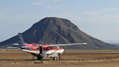 A red and white airplane is parked on a dirt landing strip with a large, dark mountain in the background under a clear sky.