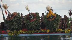 A group of men in traditional masks and costumes are on a decorated boat, possibly for a performance or festival near a body of water.