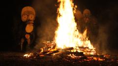 A bonfire burns brightly in the dark, flanked by blurry figures in traditional masks and costumes, likely part of a cultural ceremony.