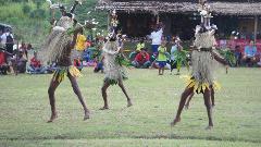 Three people in traditional costumes dance on a grassy field in front of a crowd of people.