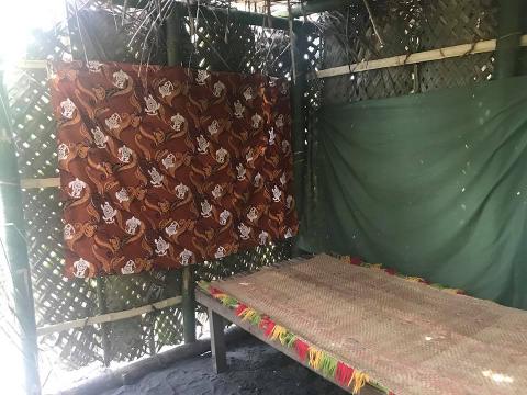 An indoor shot shows a brown fabric with turtle designs, a bed with a woven mat and colorful tassels, and a green sheet in a room made of bamboo and woven material.