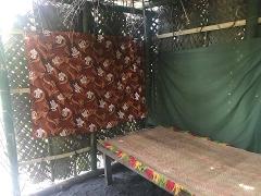 An indoor shot shows a brown fabric with turtle designs, a bed with a woven mat and colorful tassels, and a green sheet in a room made of bamboo and woven material.