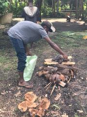 A man in a cap and jeans tends to a fire made of wood and coconut husks, while another man stands in the background in a lush, natural setting.