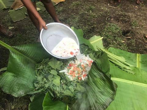 A person is pouring ingredients from a silver bowl onto a pile of green leafy vegetables and orange chunks, all resting on large banana leaves.