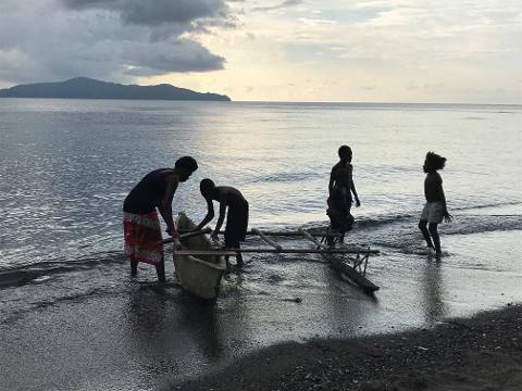 People standing in the water, pulling a small boat to shore. There are mountains in the background and clouds in the sky.