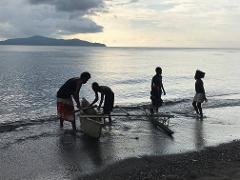 People standing in the water, pulling a small boat to shore. There are mountains in the background and clouds in the sky.