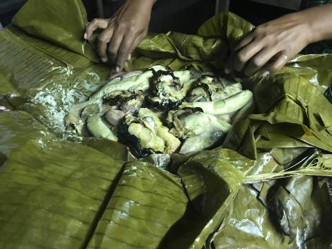 A meal wrapped in banana leaves is being prepared or served. The dish contains pieces of meat, possibly chicken, and other ingredients. Two hands are visible holding or arranging the banana leaves.