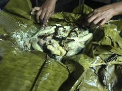 A meal wrapped in banana leaves is being prepared or served. The dish contains pieces of meat, possibly chicken, and other ingredients. Two hands are visible holding or arranging the banana leaves.
