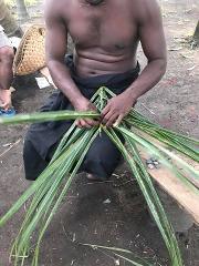 A shirtless man in Rabaul is weaving a craft with palm leaves while sitting outside.