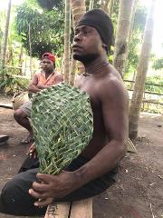 A shirtless man with a black beanie is sitting down while holding a woven fan, with another man sitting in the background.