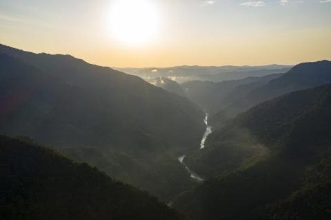 A sunlit river winds through a deep, mountainous valley, with misty layers of hills fading into the distance.