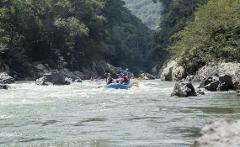 A group of people are rafting down a river with green trees and rocky banks on either side.