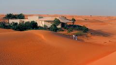 A couple walks in the desert near a resort with a large tent-like structure.