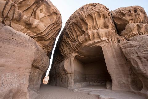 A low-angle shot captures a rock-cut tomb nestled between massive sandstone formations, showcasing ancient architectural craftsmanship against the backdrop of a desert landscape.