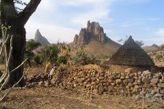 A village scene features huts with thatched roofs behind a stone wall, against a backdrop of striking rock formations and clear sky.