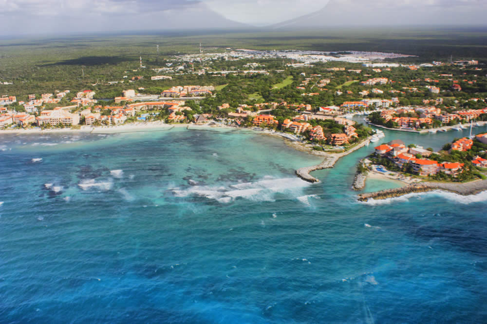 Aerial view of a tropical shoreline with turquoise waters, white sand beaches, orange-roofed buildings, and lush greenery under a partly cloudy sky.