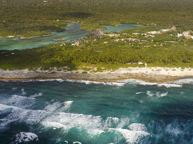 Riviera Maya Airplane Tours Aerial view of the Riviera Maya coastline showing turquoise ocean waves crashing against a rocky beach with lush green vegetation and resort buildings inland.