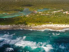 Riviera Maya Airplane Tours Aerial view of the Riviera Maya coastline showing turquoise ocean waves crashing against a rocky beach with lush green vegetation and resort buildings inland.