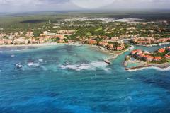 Riviera Maya Airplane Tours Aerial view of a tropical shoreline with turquoise waters, white sand beaches, orange-roofed buildings, and lush greenery under a partly cloudy sky.