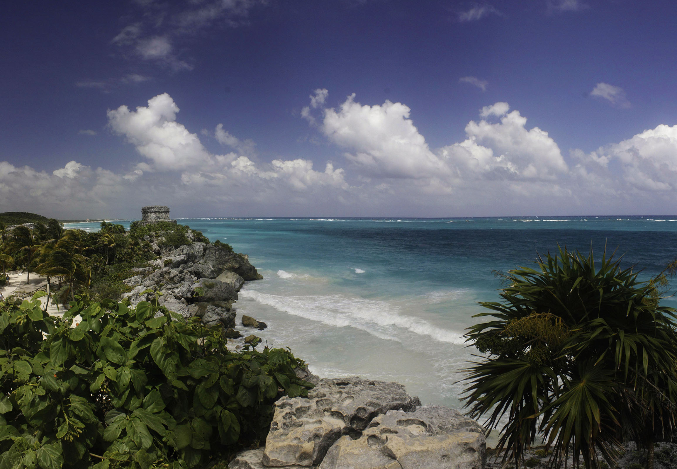 Riviera Maya Airplane Tours A beautiful coastal landscape of Riviera Maya, Mexico, featuring turquoise waters, white sandy beaches, a Mayan ruin atop a rocky cliff, and lush tropical vegetation under a partly cloudy sky.