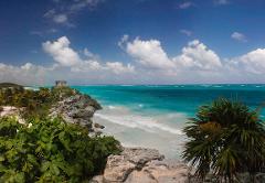 Riviera Maya Airplane Tours A beautiful coastal landscape of Riviera Maya, Mexico, featuring turquoise waters, white sandy beaches, a Mayan ruin atop a rocky cliff, and lush tropical vegetation under a partly cloudy sky.