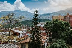 A scenic cityscape with buildings, trees, and mountains under a cloudy blue sky.