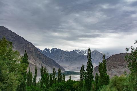 A landscape of majestic mountains under a cloudy sky, framed by lush green trees.