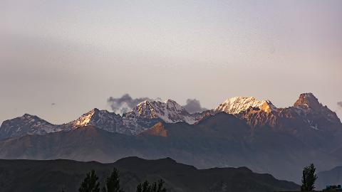 A panoramic view of snow-capped mountains at sunset, their peaks catching the golden light against a pale sky, with dark silhouettes of lower hills and trees in the foreground.