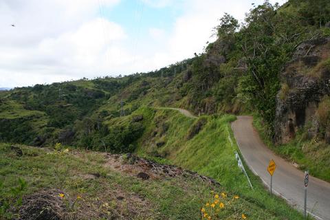 A winding road cuts through a lush, green hillside, with power lines visible in the distance.